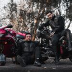 Dwóch motocyklistów w kombinezonach pozuje przy motocyklach na zewnątrz