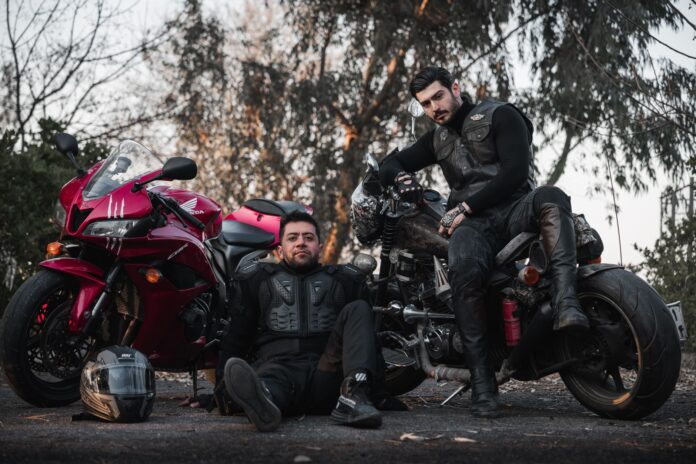 Dwóch motocyklistów w kombinezonach pozuje przy motocyklach na zewnątrz Dwóch motocyklistów w kombinezonach pozuje przy motocyklach na zewnątrz